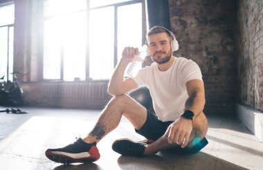 Spor giyim ve kulaklık giyen, modern çatı katında otururken ve plastik şişeden su içerken şarkı dinleyen, pozitif sakallı beyaz erkek sporcu.