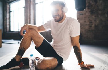 Aktivite kıyafetli arka ışıklandırmalı erkek sporcu modern ferah fitness stüdyosundaki eğitimden sonra yerde dinlenirken kulaklıkla müzik dinliyor ve gülümsüyor.