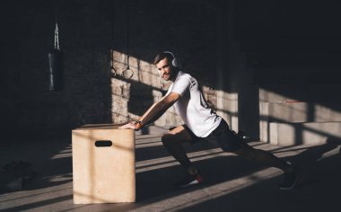 Spor giyim ve kablosuz kulaklıklı sakallı bir erkek sporcu spor salonunda ağır plyometrik kutuları taşırken kameraya bakıyor.