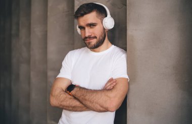 Beyaz tişörtlü, kulaklıklı, kolları çaprazlanmış, kirli gri duvarın yanında durup iyi müzik dinleyen kendine güvenen beyaz sakallı beyaz adam.