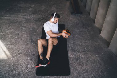 Spor kıyafetleri ve kulaklıklar takan yüksek açılı bir sporcu siyah paspasın ve antrenman bacaklarının ve ağırlık topuyla karın kaslarının üzerinde oturuyor.