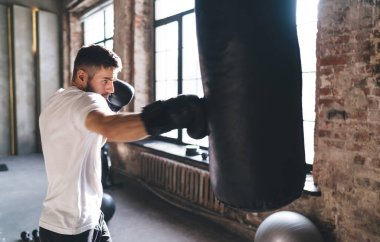 Boks eldivenli erkek sporcunun spor salonundaki konsantrasyon gücünü ve konsantrasyonu arttırmak için antrenman sırasında kum torbasıyla boks çalışmasının yan görüntüsü.