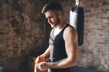 Spor salonunda boks antrenmanına hazırlanan güçlü erkek dövüşçülerin yan görüntüsü ve el bağlarını yırtılmaktan korumak için bantla sarma.