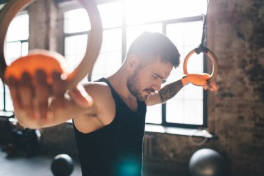 Kafası aşağıda ve elleri jimnastik halkalarına sarılı duran güçlü kaslı erkek sporcunun yan görüntüsü. Güneş ışığında antrenman yaparken spor salonunu yansıtıyor.
