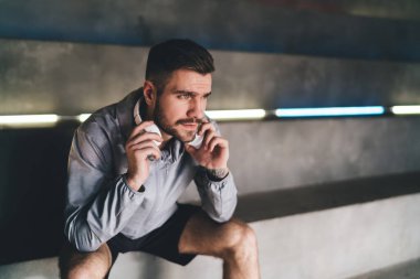 Düşünceli sakallı erkek sporcu elinde kulaklıklarla beton merdivenlerde oturuyor ve spor salonunda dinlenip zaman geçirirken başka tarafa bakıyor.