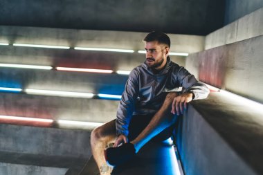 Spor ceketli, düşünceli, sakallı bir erkek modern spor salonunun ışıklı merdivenlerinde oturup yoğun bir eğitimden sonra dinlenirken yüzünü çeviriyor.