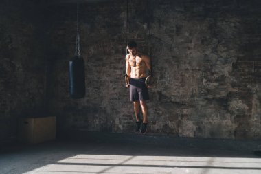Çatı katı tarzı güneşli spor salonunda spor yaparken çıplak vücutlu beyaz erkek sporcu jimnastik yüzükleri üzerine egzersiz yapıyor.