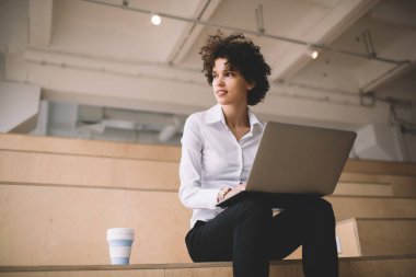 Milenyum Kafkas kızı dizüstü bilgisayar kullanırken gözlerini kaçırıyor. Modern başarılı kadın kavramı. Genç, yoğun kıvırcık kadın, çalışma odasında, ahşap bankta kahve fincanıyla oturuyor.