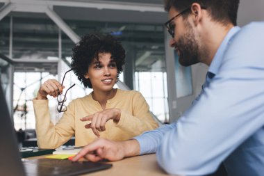İş kadını, kendisine bakan erkek arkadaşını dizüstü bilgisayarla işaret ediyor. İş birliği ve takım çalışması. Genç Kafkasyalı milenyum insanları ofiste masa başında. Modern başarılı insanlar