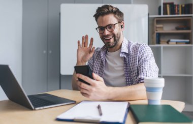 Milenyum Kafkas işadamı akıllı telefondan video görüşmesi sırasında el sallıyor. Modern başarılı adam kavramı. Gözlüklü ve kulaklıklı genç adam açık uzay ofisindeki masada oturuyor.