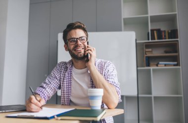 Milenyum Kafkas işadamı akıllı telefonla konuşuyor, panoya yazı yazıyor ve başka tarafa bakıyor. Modern başarılı adam kavramı. Gözlüklü genç adam açık uzay ofisinde masada oturuyor.