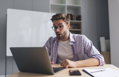 Bin yıllık Kafkas iş adamı iş sırasında dizüstü bilgisayardan bir şey izliyor. Modern başarılı adam kavramı. Gözlüklü, ağzı açık, uzay ofisinde masada oturan heyecanlı genç adam.