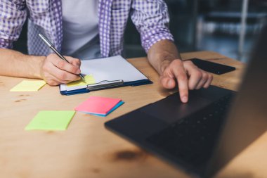 Milenyum işadamının iş sırasında dizüstü bilgisayar kullanıp panoya yazı yazarken kısmi görüntüsü. Modern başarılı adam kavramı. Ofisinde bilgisayarını açmış ahşap masadaki adam.