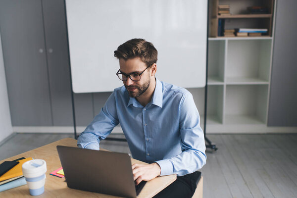 Millennial caucasian businessman typing on laptop computer during work. Concept of modern successful man. Young concentrated guy wearing glasses and formal suit sitting at desk in spacious office