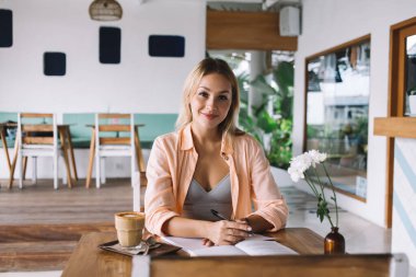 Gülümseyen genç bir kadın elinde bir bardak kahveyle masaya oturmuş kitap okurken ve kafede boş vakit geçirirken kalemle notlar yazarken kameraya bakıyor.