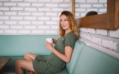 Kanepede elinde bir fincan sıcak içecekle oturan ve kafeteryada, duvardaki aynayla dinlenirken kameraya bakan neşeli genç bir kadın.