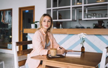 Genç bir bayanın elindeki kalemle defterden notlar alırken ve kafede bir bardak kahveyle masada otururken yan bakışları.