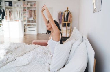 Gecelik giymiş genç bir bayanın rahat bir yatak odasında yatarken kollarını açması ve evdeki sabahın tadını çıkarması.