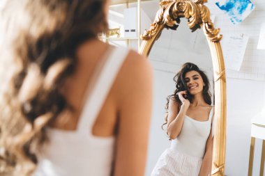 Pijamalı, uzun dalgalı saçlı, aynadaki yansımasına hayran olan ve sabah yatak odasında uyandıktan sonra gülümseyen zayıf kadının arka görünüşü.