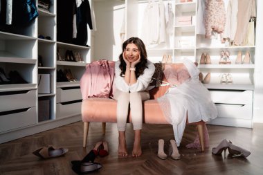 Pembe kanepede oturan ve giysi odasındaki dolabın yanındaki ayakkabıları seçen zarif genç bir kadın.