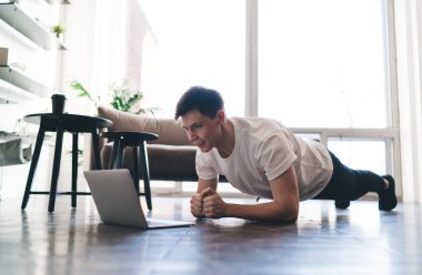 Dizüstü bilgisayara bakarken ön kol tahtası egzersizi yapan genç adamın yan görüntüsü ve evdeki oturma odasında boş zamanlarında izlenen videodaki adımlar.