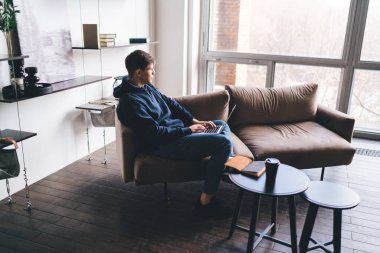 Mavi gömlekli ve kot pantolonlu genç bir adamın yan görünüşü. Kanepede oturup pencereden dışarı bakarken evdeki oturma odasında uzaktan proje üzerinde çalışıyor.