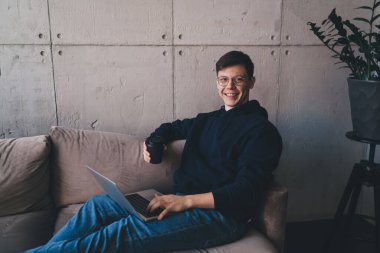 Gözlüklü genç adam bilgisayar kullanırken saksı bitkisinin yanındaki koltukta oturuyor.