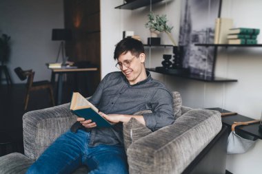 Gözlüklü genç adam rahat oturma odasındaki kanepede dinlenirken kitap okuyor ve evde mutlu zaman geçiriyor.