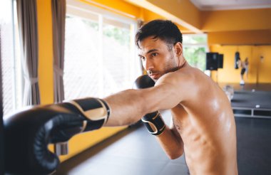 Boks eldivenleri giymiş güçlü bir adamın yan görünüşü. Gün ışığında pencerelerin yanında duran ve perdelerle kum torbasını yumruklayan. Gözünü kaçıran ve bulanık spor salonunda antrenman yapan.