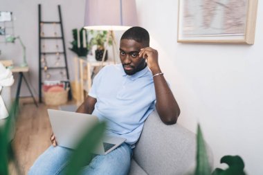 Ciddi bir Afro-Amerikan erkek serbest çalışan, gündelik kıyafetlerle kanepede oturmuş dizüstü bilgisayarla çalışırken başka tarafa bakıyor.