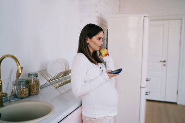 Mutfakta durup cep telefonuna göz atan sakin hamile bir kadının üçüncü üç aylık hamilelik dönemini doğum için hazırlanırken sağlıklı bir atıştırmalık yemesi.