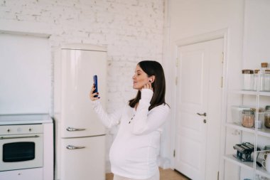 Müstakbel, mutlu, siyah saçlı, beyaz elbiseli, cep telefonuyla video görüşmesi yapan ve evde, ferah, aydınlık bir mutfakta duran annenin yan görüntüsü.