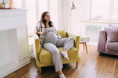 Sakin, hamile, siyah saçlı, günlük kıyafetler giyen, modern ışıklı oturma odasındaki rahat koltukta uzanan bir anne.