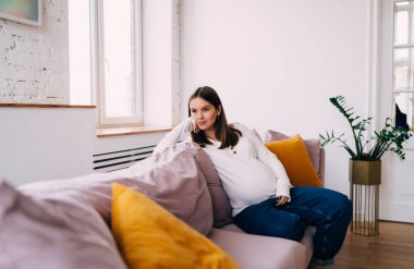 Bebek bekleyen genç bir kadın rahat bir kanepede dinleniyor. Yastıklar yanaklarında. Düşünceli bir şekilde geleceği düşlüyor.