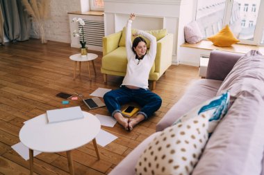 Uzaktan kumandalı bir projede çalışırken ellerini kaldırıp gülümseyen ve dizüstü bilgisayarlı akıllı telefon ve kağıtlarla yerde dinlenen düşünceli, genç ve mutlu bir kadın serbest yazar.