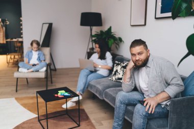 Üzücü sakallı baba, hafta sonu evde birlikte dizüstü bilgisayar ve cep telefonu kullanırken koltuğa yaslanıp elini kolunu sallayarak oturuyor.