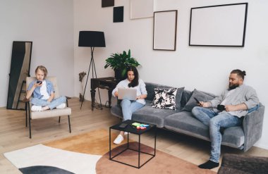 Bütün vücudu sakallı bir adam, günlük kıyafetleriyle koltukta oturuyor, akıllı telefonuyla bir kadınla mesajlaşıyor, dizüstü bilgisayar kullanıyor, kızı da sosyal medyada geziniyor.