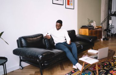 Haftasonu rahat bir kanepede cep telefonuyla dinlenirken bir fincan sıcak içecek mesajlarıyla tam boy Afro-Amerikan erkek.