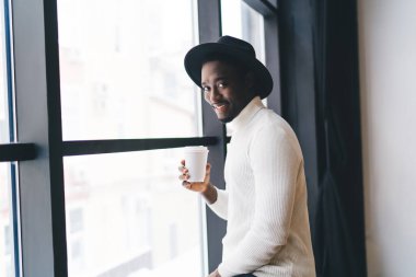Beyaz balıkçı yakalı, elinde kağıt bardak taze içecekle, haftasonu boyunca modern kafeteryanın penceresinin yanında kameraya bakıp boş zamanın tadını çıkaran pozitif siyah bir adam.