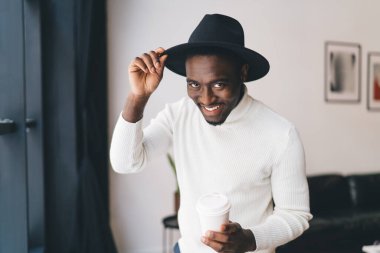 Beyaz balıkçı yakalı genç Afro-Amerikalı adam siyah şapkaya dokunuyor ve molada sıcak içecek içerken kameraya bakıyor.