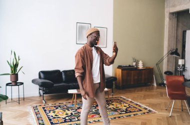 Şapkalı ve gözlüklü Afro-Amerikan bir erkeğin TWS kulaklıklarıyla yürürken ve cep telefonuyla selfie çekerken görüntüsü. Ekrana bakarken ve evde müzik dinleyerek zaman geçirirken.