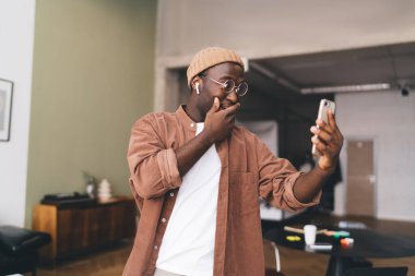 Dilsiz, genç, gözlüklü ve kulaklıklı Afrikalı bir erkek. Oturma odasındaki çevrimiçi video görüşmesi sırasında ayakta durup cep telefonunun ekranına bakarken eliyle ağzını kapatıyor.