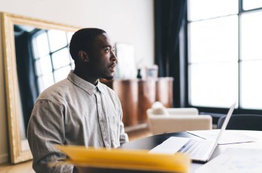 Afrika kökenli Amerikalı bir erkek, elinde bilgisayarla masada oturup evdeki oturma odasında uzaktan kumandayla düşünürken yüzünü çeviriyor.