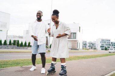 Hafta sonları asfalt kaldırımda yürürken kız arkadaşının kaymasına ve gülmesine yardım eden sıradan giysili Afrikalı Amerikalı erkek arkadaş.