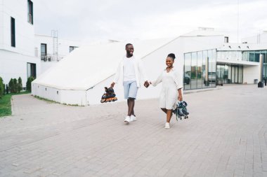 Modern şehir mahallesinde kaldırımda yürürken hafif giysiler içinde el ele tutuşan ve paten taşıyan genç siyahi kadın ve erkekler.