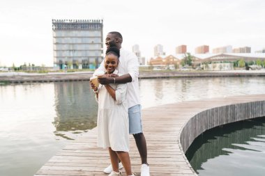 Genç Afrikalı erkek ve kadın ellerinde kahve fincanlarıyla kentte romantik bir buluşma için asfalt kaldırıma çıkarken kucaklaşıyorlar.