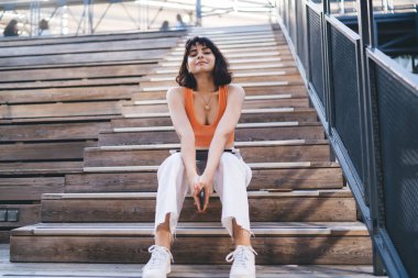 Genç ve modern bir kadın vücudu var. Kısa kahverengi saçlı. Üstü açık, beyaz kot pantolonlu ve merdivenlerde oturmuş kameraya bakıyor.