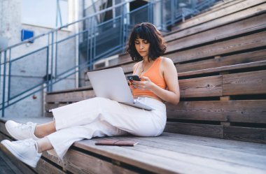 Normal kıyafetleri ve spor ayakkabıları içinde, dizüstü bilgisayarla ahşap merdivenlerde oturan ve uzaktan çalışan akıllı telefondan mesaj atan bir sürü konsantre genç kadın.