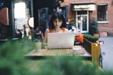Siyah saçlı genç bayan dijital göçebe, tahta masada kahve bardağıyla oturuyor ve sokak kafesinde dizüstü bilgisayar kullanarak proje üzerinde çalışıyor.