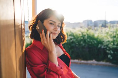 Kırmızı günlük kıyafetli genç bir kadın kameraya bakıyor ve ahşap duvara yaslanırken cep telefonuyla konuşuyor.